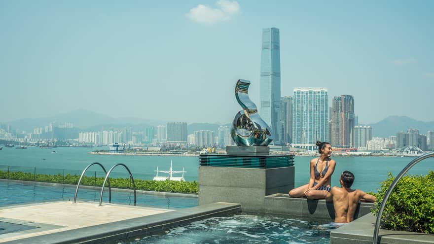 A couple in swimwear sit on the edge of an infinity pool with a harbor view, overlooking a city skyline, modern sculpture and mountains under a clear sky.