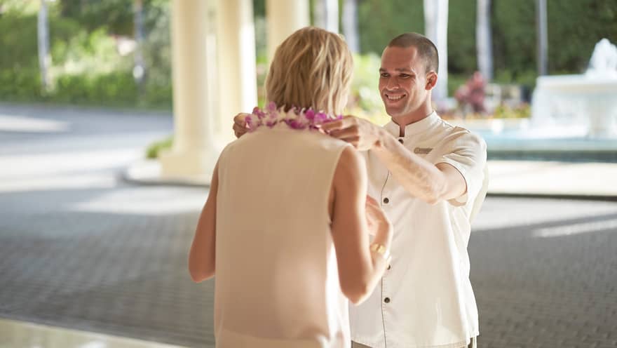Hotel staff member smiling while placing a purple flower lei on a guest during arrival