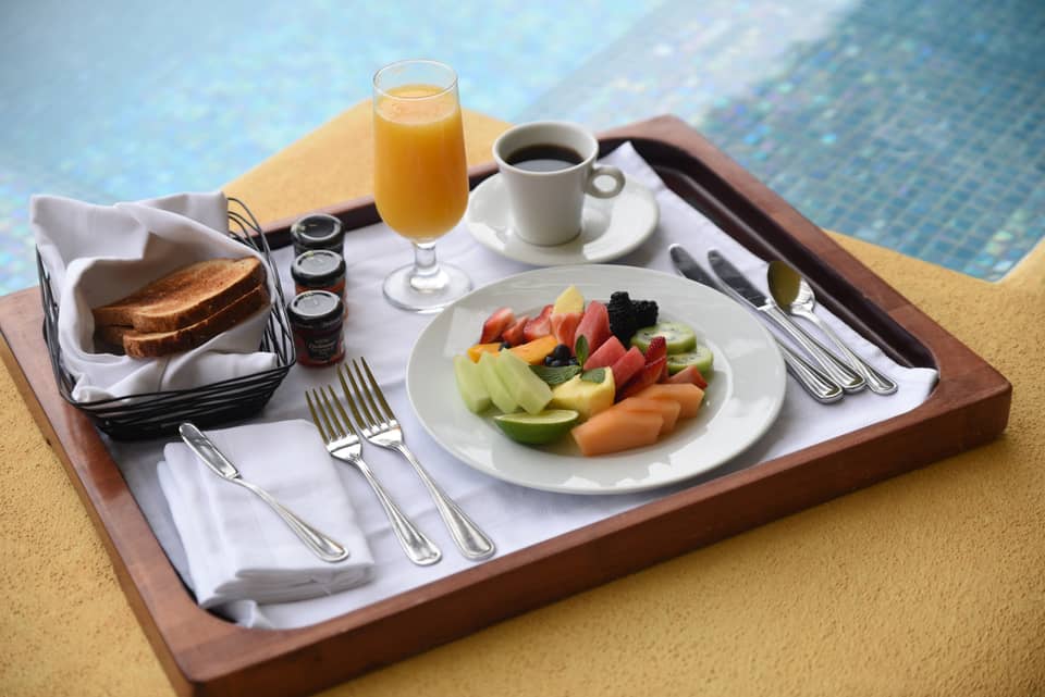 Fresh fruit slices on plate, coffee, orange juice, toast on wood tray by pool