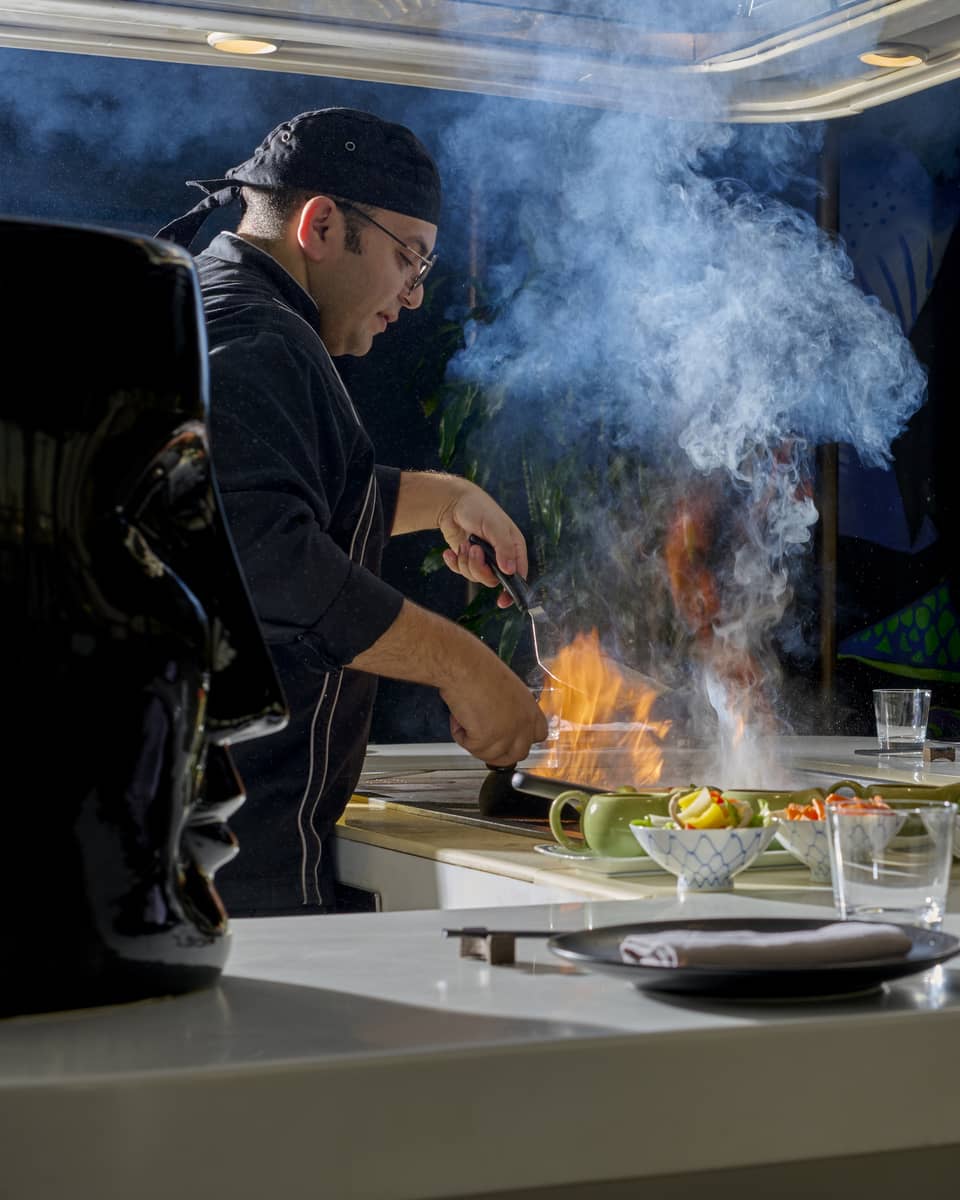 Chef in black coat cooks dish over open flame in show kitchen