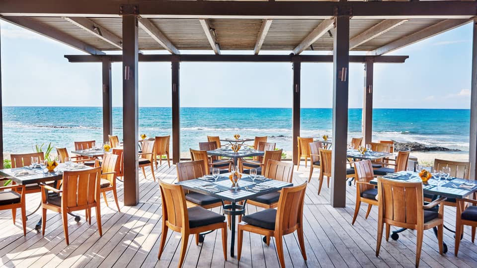 The Ulu Ocean Grill and Sushi Lounge is ready for guests with square tables set with menus and clear glassware, and overlooking the ocean from the covered wooden terrace.