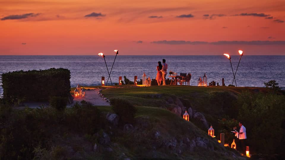 A couple stands on The Rock outpost at dusk, overlooking the ocean, with private dining table with lanterns and torches