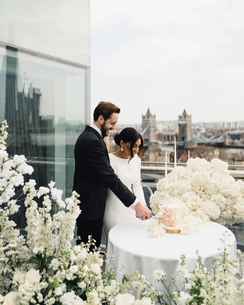 A couple cutting the cake together on a terrace with a view of the city skyline, surrounded by white flowers