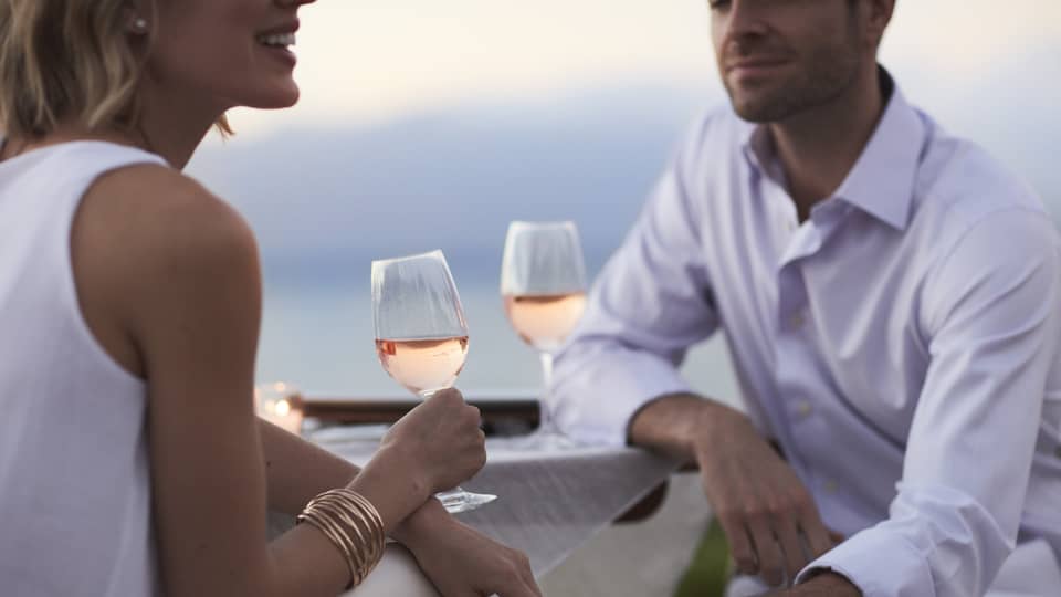 Close-up of woman and man in white, sitting and holding glasses of rose wine at dusk