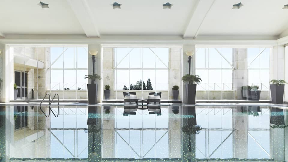 Indoor swimming pool reflecting light of three tall windows, white walls and pool chairs