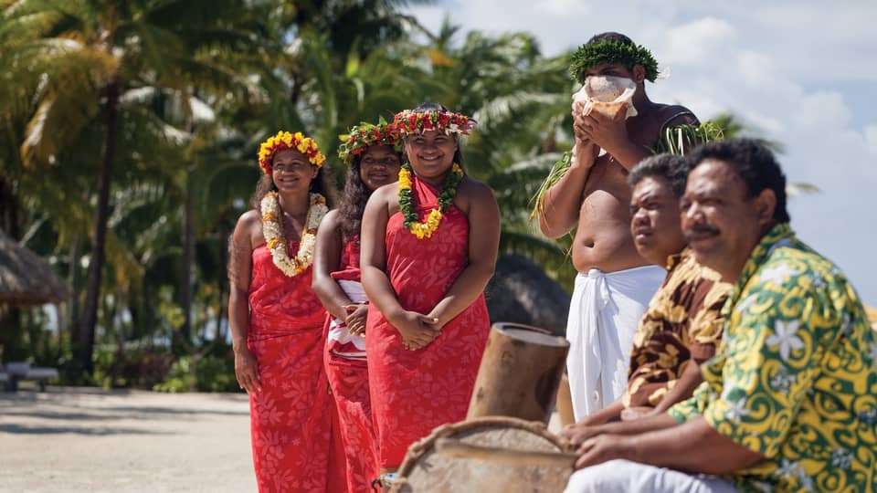 Group of people in traditional attire with leis and a conch shell, standing on a beach
