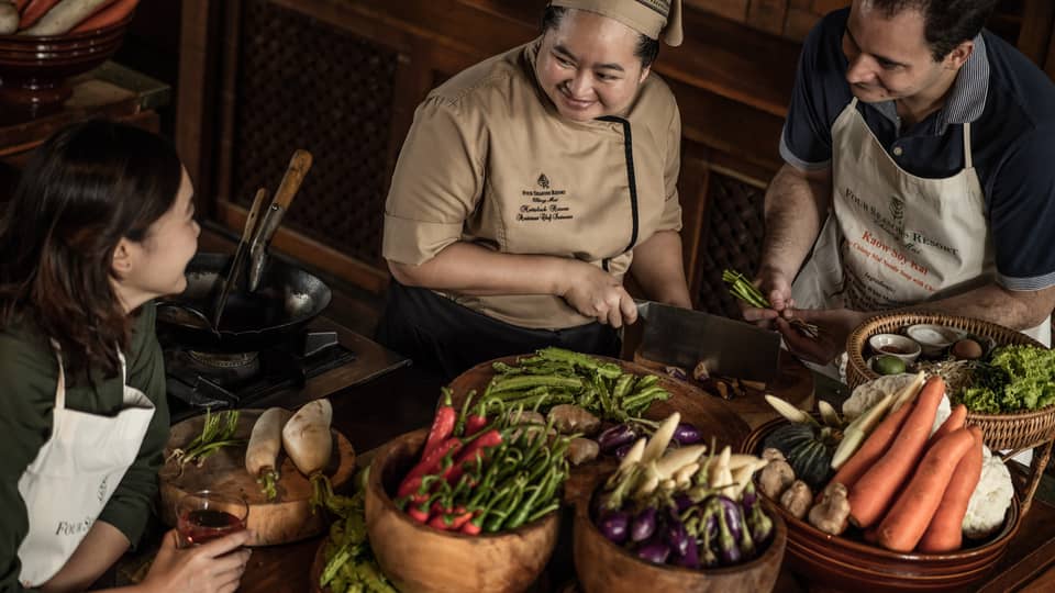 A chef teaching two participants in a cooking class, surrounded by fresh vegetables and ingredients in a rustic kitchen setting.