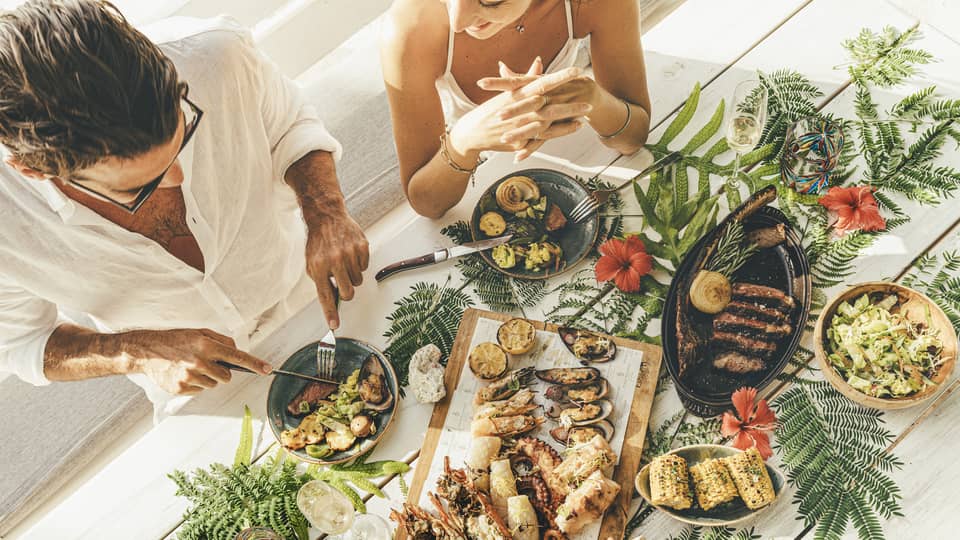 A couple at a table of seafood canapés, grilled meat, corn and champagne flutes, adorned with palm fronds and red hibiscus.