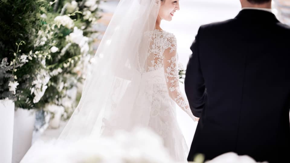 A bride in a white, lace, long sleeve gown smiles as she walks down the aisle with her groom. They are surrounded by artfully arranged white flowers.