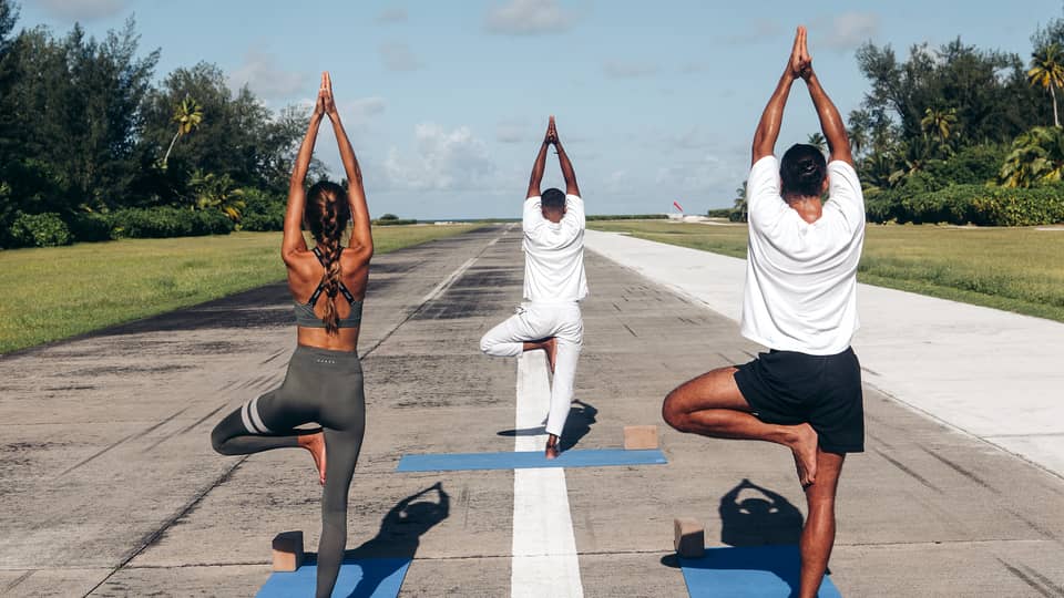 Rear view of three people practicing yoga in tree pose on an airport runway framed by lush greenery, under a clear blue sky.