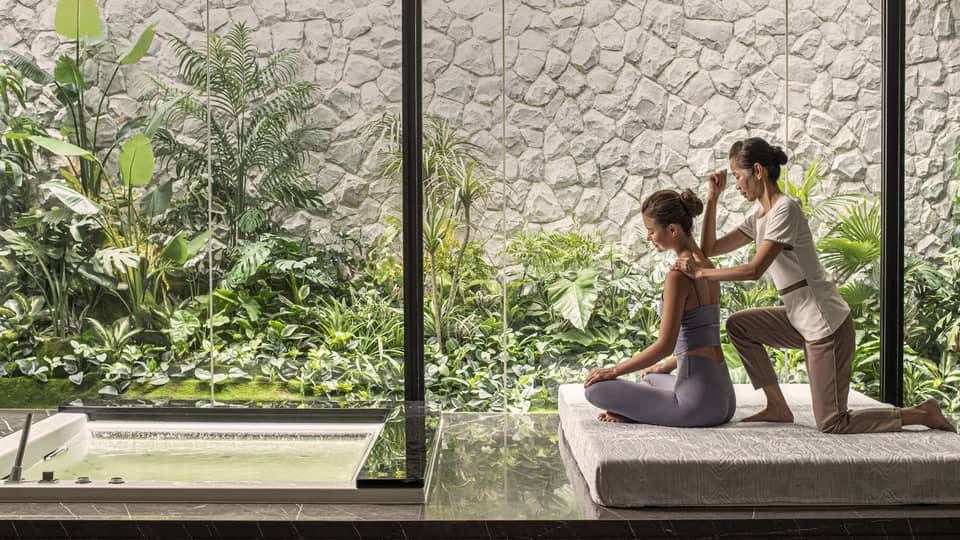Person receiving a massage in a peaceful spa setting with large windows overlooking lush green plants and a small water feature.