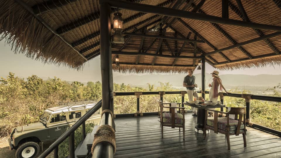 A couple sitting on a wooden deck overlooking green brush, Land Rover sitting nearby