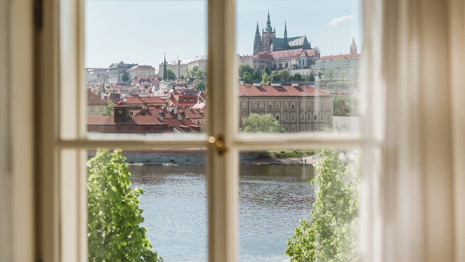 View through a window of a historic city with red rooftops and a cathedral in the distance, overlooking a river and green trees under a clear blue sky.