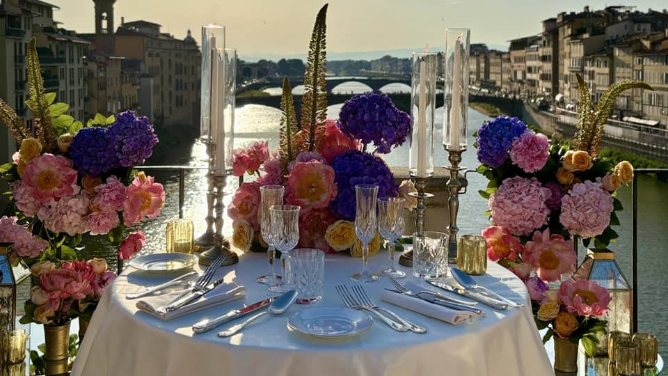 An elegantly set outdoor dining table overlooking a a river, with a sunny sky in the background.