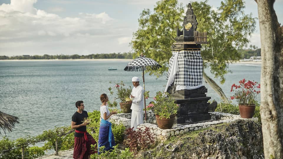 High Priest Aji Ngurah stands by shrine with black-and-white checkered umbrella, cloth, as man and woman make offering