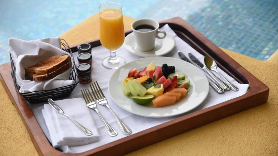 Fresh fruit slices on plate, coffee, orange juice, toast on wood tray by pool