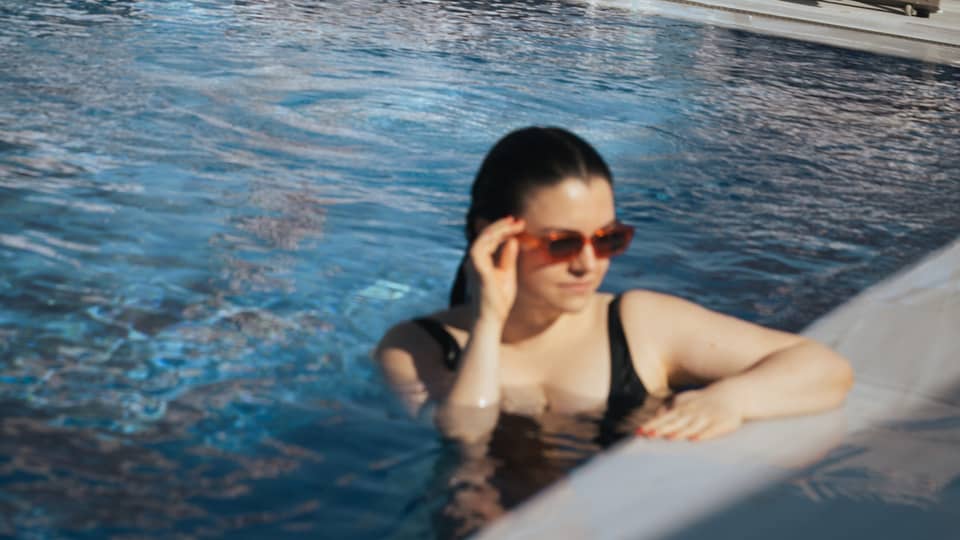 A person in a bathing suit and sunglasses at the edge of a clear blue pool. Lounge chairs, umbrellas and the distant landscape are in the background