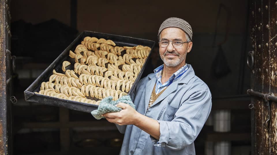 Pastry chef in casual blue coat, hats holds tray with crescent-shaped corned de gazelle