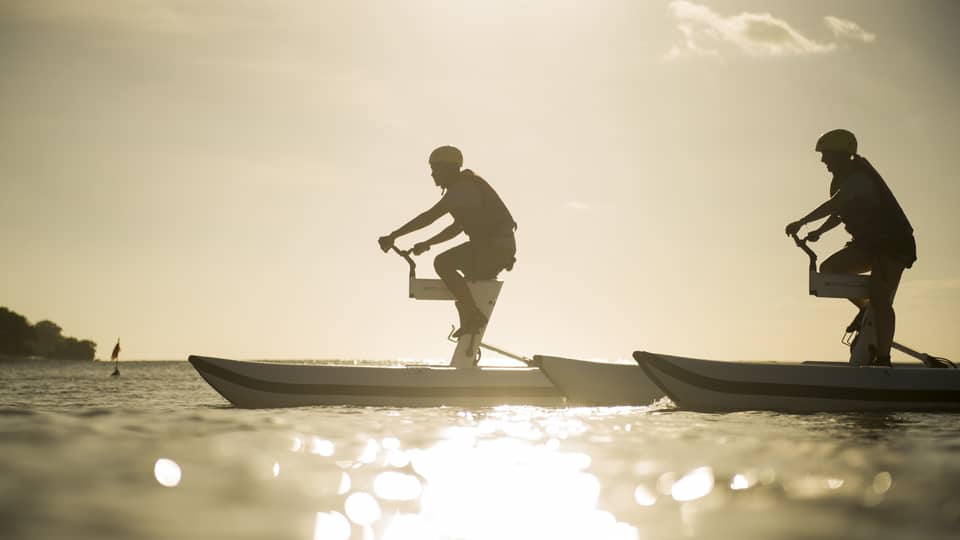 Silhouettes of two men riding aquatic bicycles on ocean at sunset