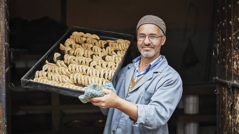 Pastry chef in casual blue coat, hats holds tray with crescent-shaped corned de gazelle
