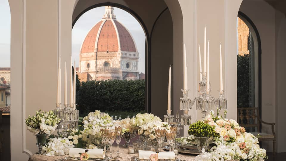 Elegant dining table set with candles, flowers, and glassware, with a large dome visible through arched windows in the background.