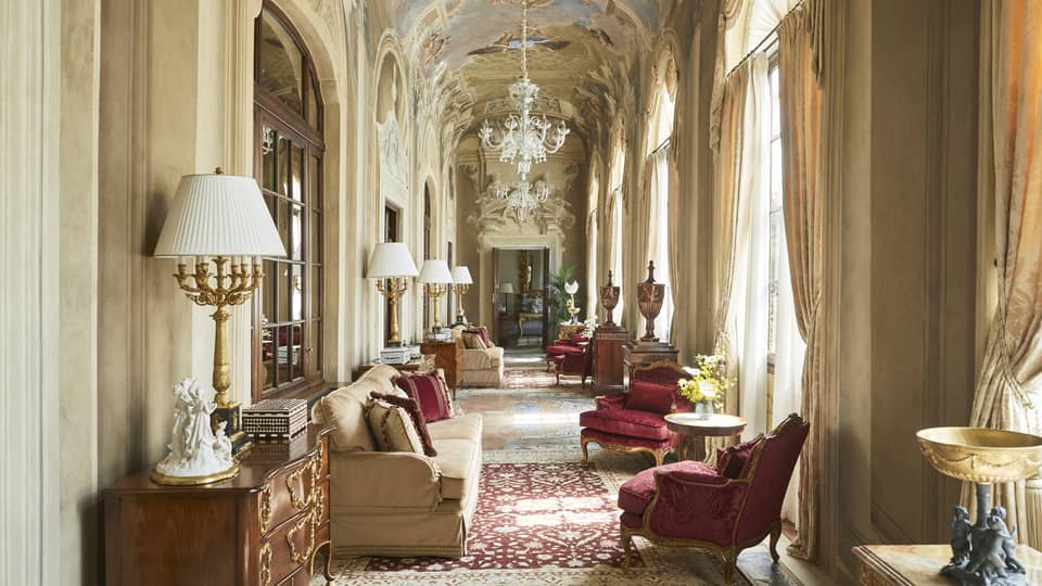 Grand, arched hallway with frescoed ceiling and chandelier, lounge seating and ruby-red arm chairs, in the Royal Suite at Four Seasons Hotel Florence