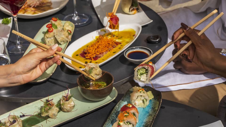 Three hands picking up sushi with chopsticks over plates of dishes
