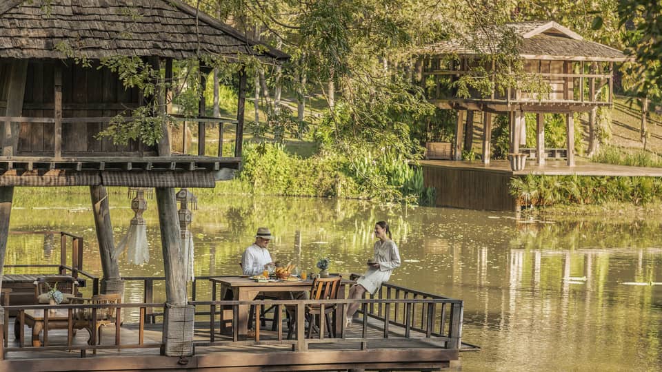 Two guests at a rustic table, on a platform in one of two wood huts on stilts perched atop a calm river bordered by greenery.