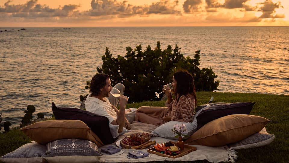 A man and woman lay on blanket and pillows during luxury picnic at sunset, next to the ocean