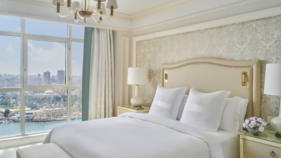 Guest bedroom with king bed, white linens and cream tufted headboard; floor-to-ceiling window with views of the River Nile