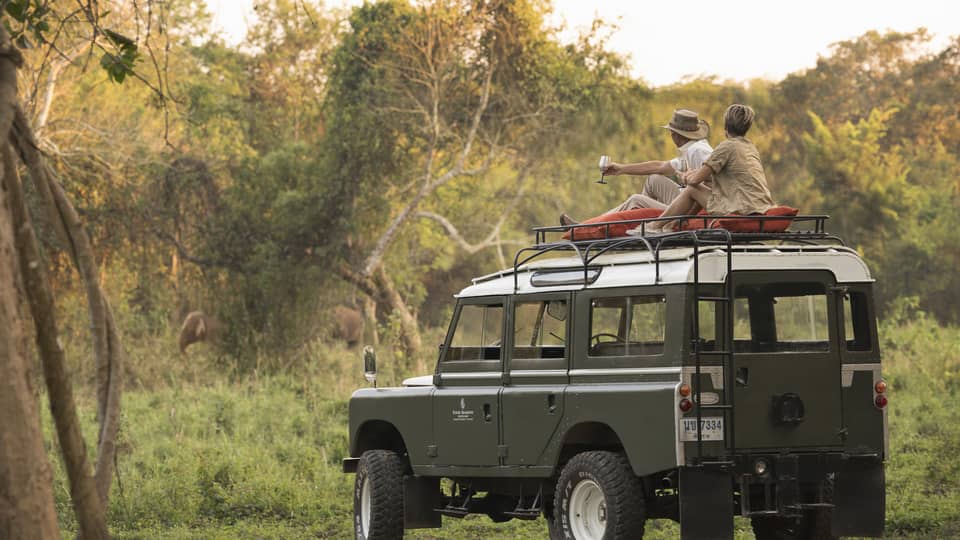 A couple sits on the roof of a Land Rover in the Bush, drinking a cocktail