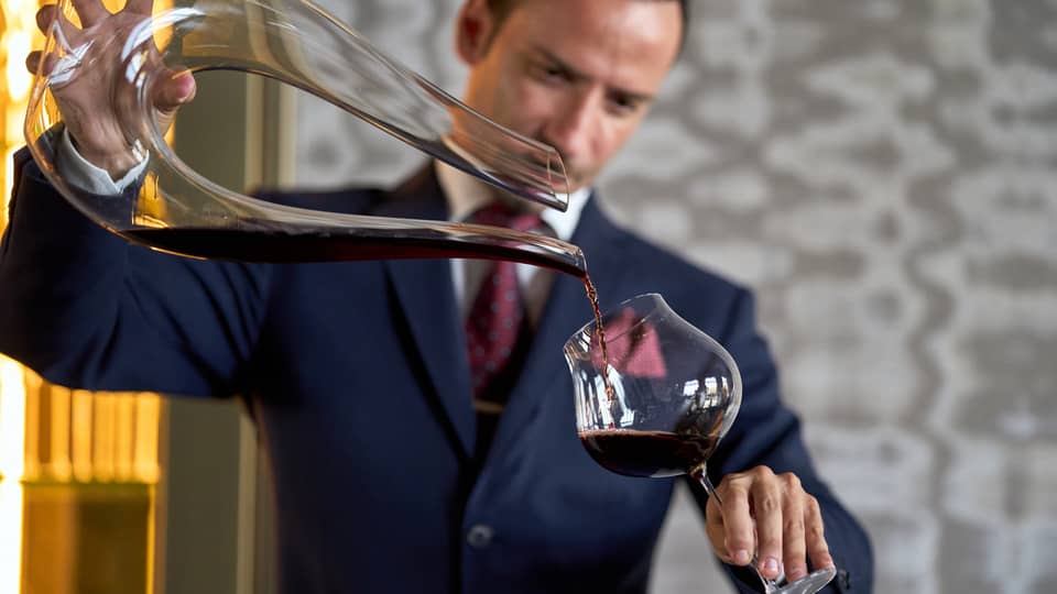 A person in formal attire carefully pouring red wine from a decanter into a glass.
