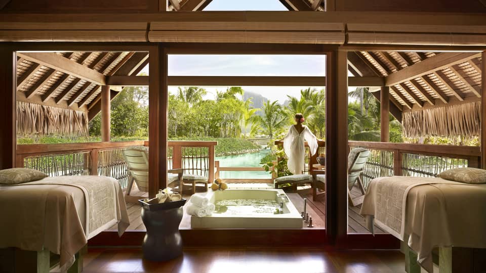 Woman wearing flowing white robe stands at edge of spa suite near plunge bath, couples massage beds