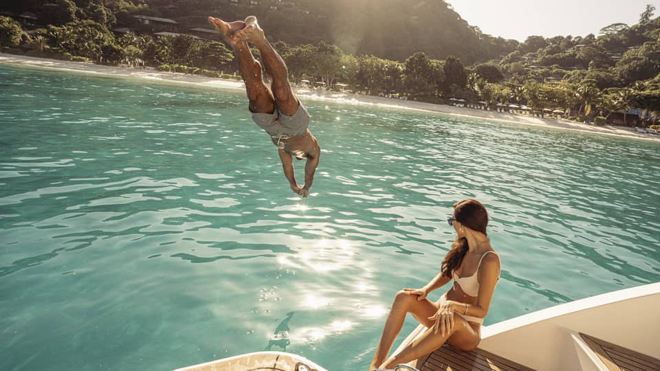 Person diving into blue-green water off a boat deck, where a person in swimsuit is sitting
