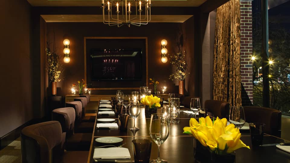 Long private dining table with plush chairs set with wine glasses, yellow flowers, candles, low lights on walls