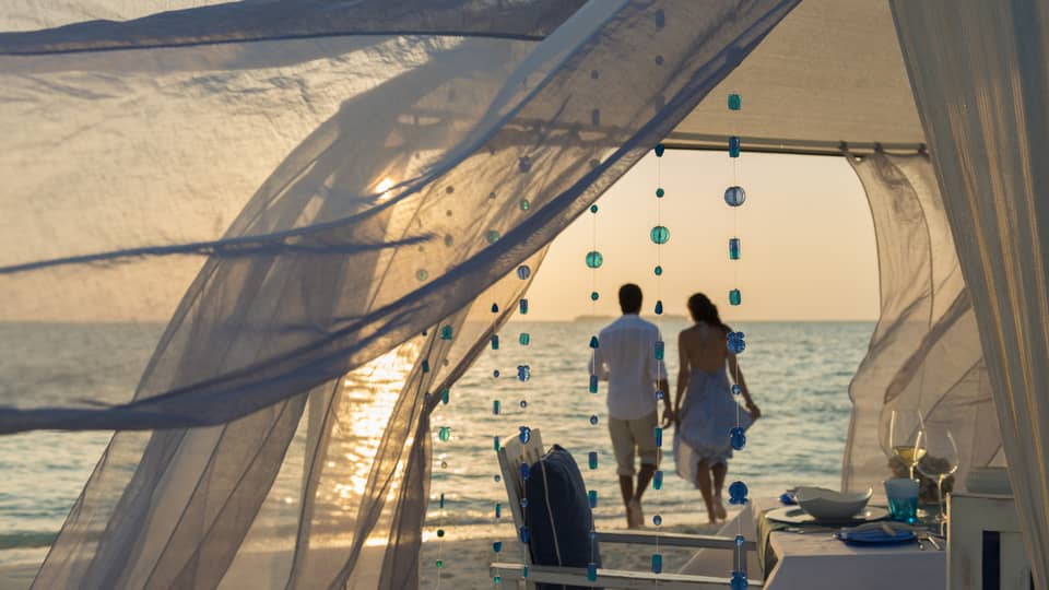 Sheer curtains blow in breeze, small blue beads hang from string, woman and man hold hands on beach in background