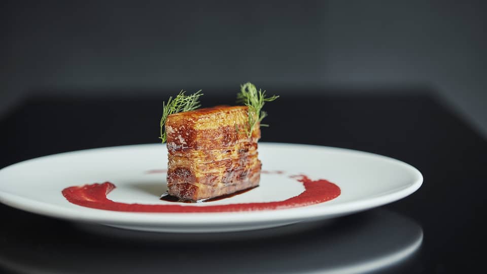 Pork belly slab on white plate with red sauce and green garnishes