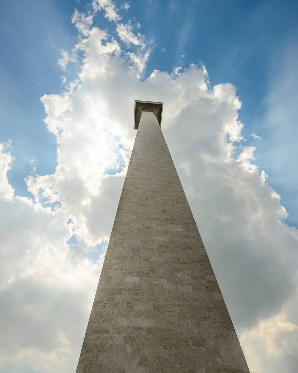 Staring up from the base of the Monas National Monument; a blue sky and the sun peeking through the clouds as a backdrop.