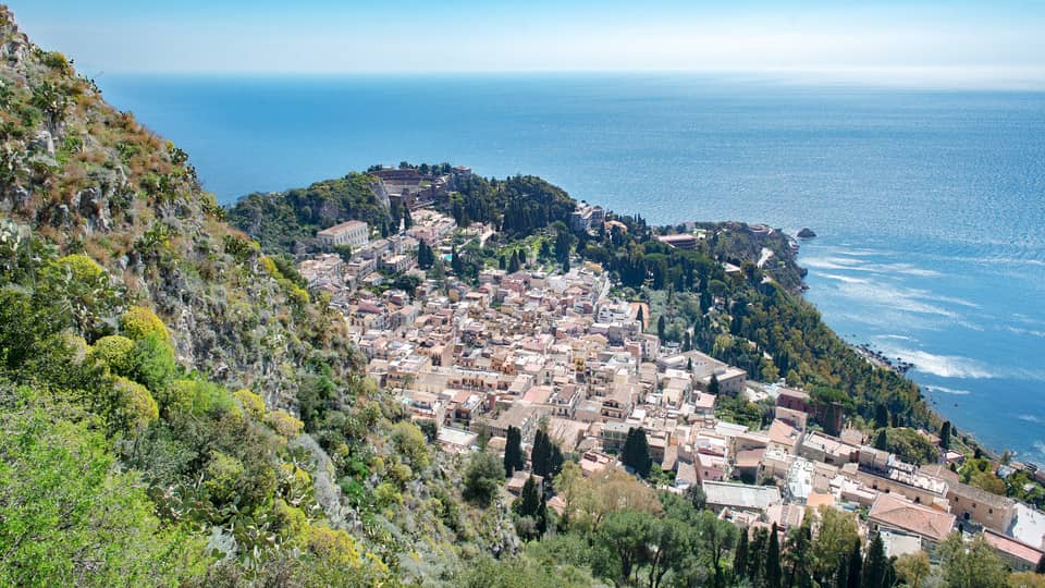 Aerial view of a coastal town with clustered buildings, lush greenery and the blue expanse of the ocean in the background.