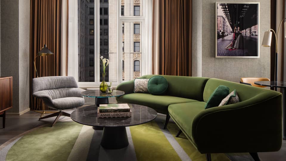 Suite living room with green sectional sofa, round coffee tables, gray armchair, wall art, and large window overlooking city buildings