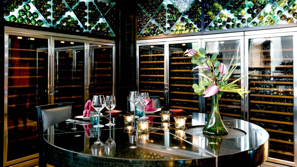 Opulent setting of goblets, candles and a tropical flower atop a mirrored table, surrounded by floor-to-ceiling wine racks.