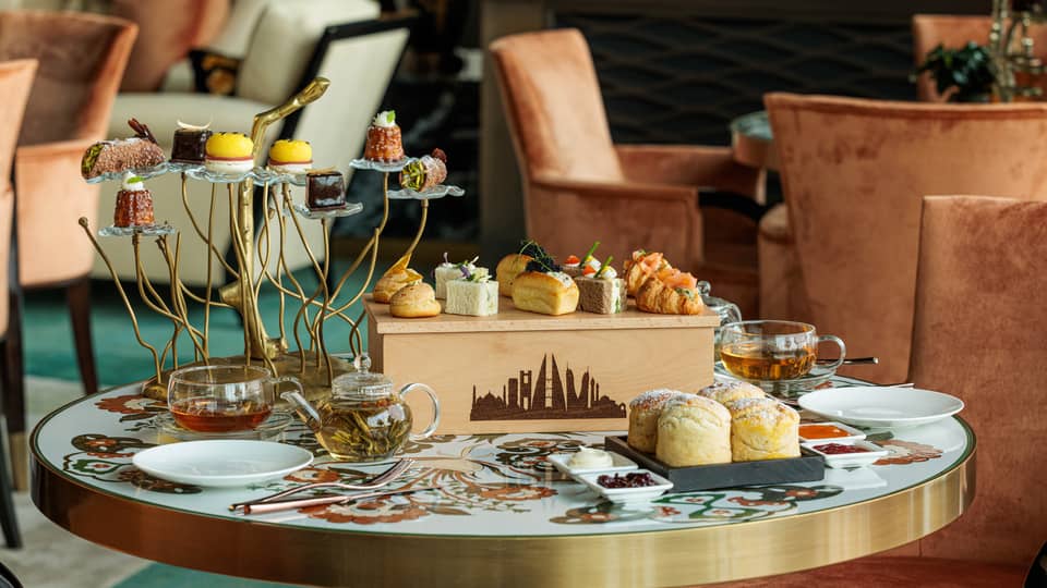Table set for afternoon tea featuring a glass teapot and cups, various pastries on a wooden box and a tree-like pastry stand with a pastry on each branch