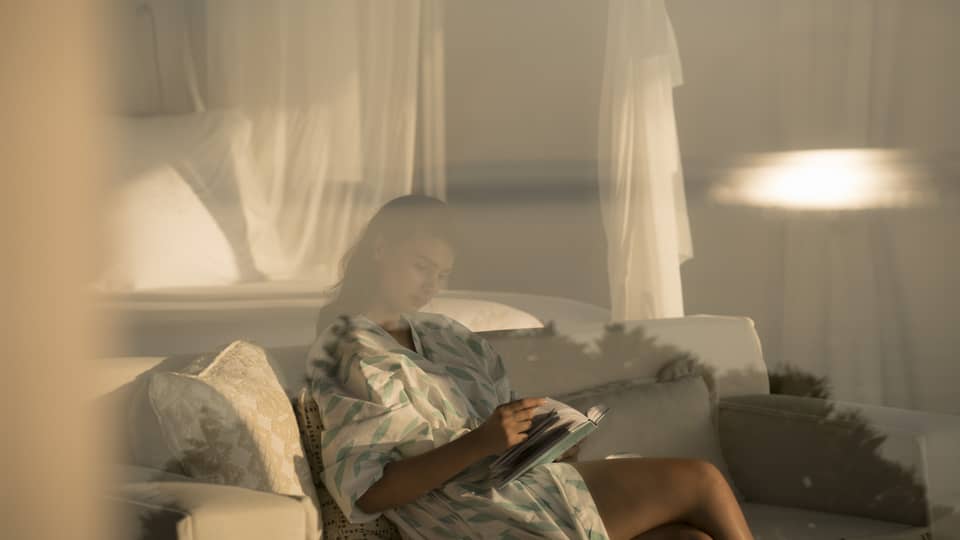 Woman in white robe with green print behind glass, reads book on white sofa