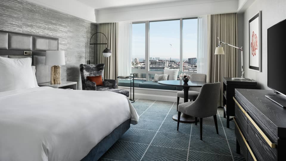 Modern hotel room with king bed, table and two upholstered grey chairs, windows overlooking San Francisco
