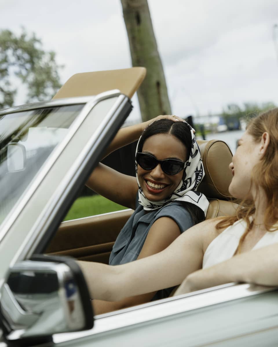 Two guests riding in a convertible car while smiling at one another.