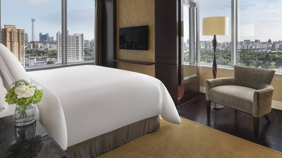 Hotel room with king bed, arm chair, wall-mounted TV and two walls of windows with river view