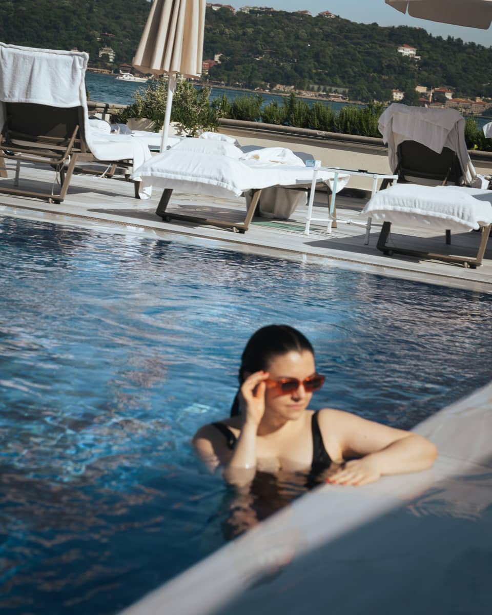 A person in a bathing suit and sunglasses at the edge of a clear blue pool. Lounge chairs, umbrellas and the distant landscape are in the background