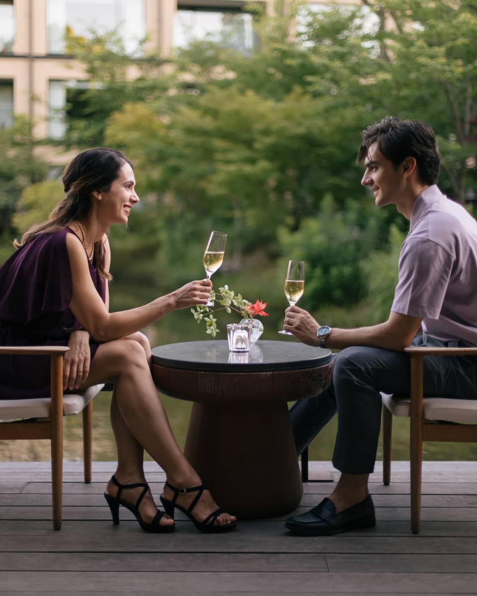 Two people sitting outdoors, toasting with champagne glasses at a small table with a flower vase