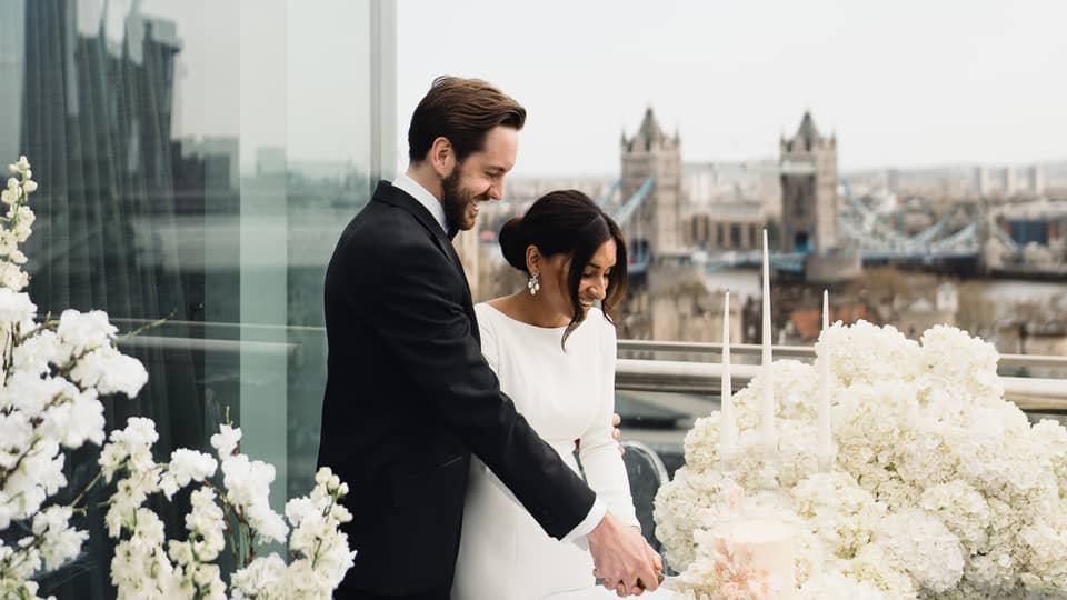 A couple cutting the cake together on a terrace with a view of the city skyline, surrounded by white flowers
