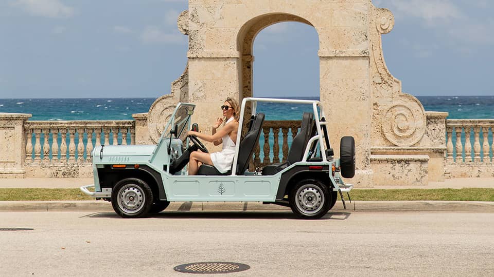 A person drives a light blue open-top vehicle past a historic clock tower by the sea. The backdrop features clear skies and the ocean.
